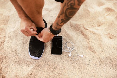 Téléchargez les photos : Runner homme attachant des lacets de chaussures de course se préparant pour courir avec smartphone et écouteurs près de lui, se préparant pour l'entraînement sur la plage, vue de dessus, maquette, espace de copie - en image libre de droit