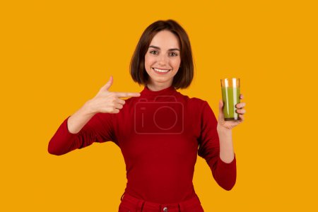 Téléchargez les photos : Joyeux joyeux jolie brune jeune femme portant une tenue décontractée rouge pointant du doigt le verre avec du jus de smoothie vert à la main et un fond de studio orange souriant. Concept de mode de vie sain - en image libre de droit