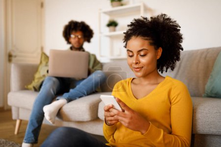 Téléchargez les photos : Black Lady Websurfing sur Smartphone tandis que son mari en utilisant ordinateur portable assis sur le canapé à la maison. Jeune couple utilisant des gadgets dans le salon moderne le week-end. Focus sélectif - en image libre de droit