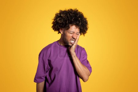 Photo for Despaired sad adult african american curly guy in violet t-shirt presses hand to cheek, suffering from toothache isolated on yellow studio background. Caries, dental problems, health care - Royalty Free Image