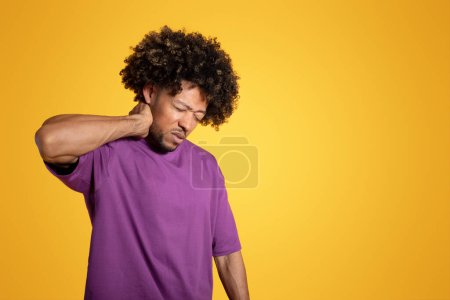 Photo for Sad adult african american curly man in purple t-shirt presses his hand to neck and suffering from muscle pain, isolated on yellow studio background. Injured, health problems - Royalty Free Image