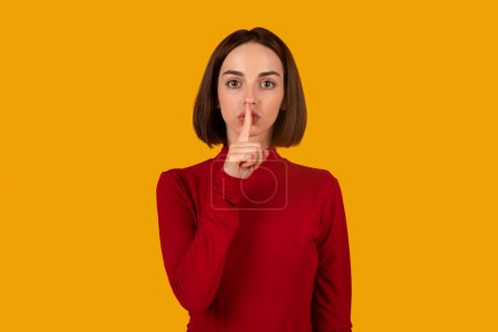 Photo for Serious mysterious brunette young woman presses index finger to lips, makes hush gesture, tells secret, asks to be quiet, wears long sleeved jumper, poses against orange background. Secrecy concept - Royalty Free Image