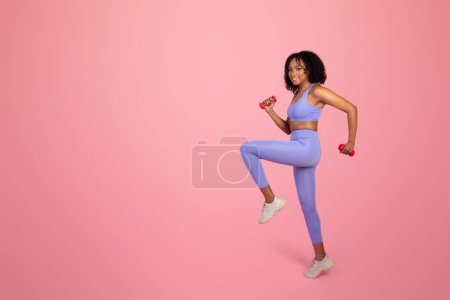 Photo for Cheerful young african american lady in sportswear doing cardio exercise with hand dumbbells, enjoy sport alone on pink studio background, full length. Fit lifestyle, ad and offer, body care - Royalty Free Image