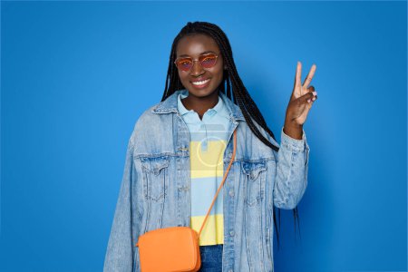 Téléchargez les photos : Mode, style. Joyeux heureux sympathique jolie jeune femme noire dans une tenue décontractée élégante et des lunettes de soleil posant sur fond de studio bleu, montrant la paix V geste et souriant à la caméra - en image libre de droit