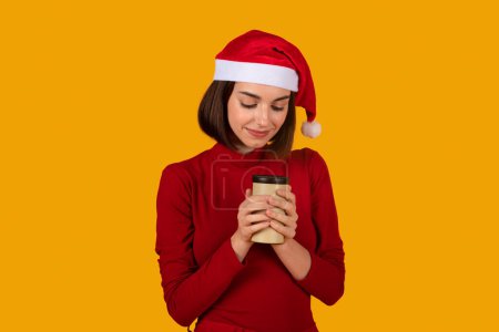 Photo for Pretty smiling young brunette woman wearing Santa hat holding paper cup coffee to go with both hands over orange studio background, warming up during cold winter days, copy space - Royalty Free Image