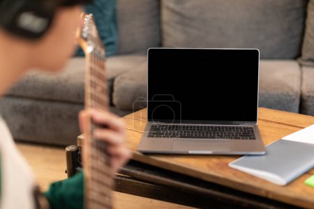 Téléchargez les photos : Technologie et musique. Homme méconnaissable jouant des accords sur guitare électrique près d'un ordinateur portable avec écran blanc à l'intérieur. Site Web musical ou publicité de logiciel en ligne. Focus sélectif sur l'ordinateur - en image libre de droit