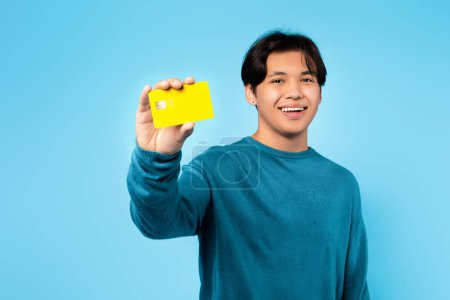 Téléchargez les photos : Un étudiant asiatique joyeux montrant de la publicité par carte de crédit Grande offre bancaire et effectuant un paiement debout sur fond bleu studio, souriant à la caméra. Publicité des services financiers - en image libre de droit