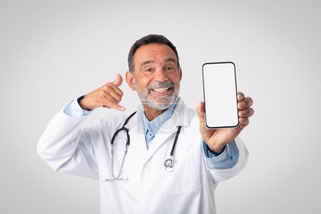 Téléchargez les photos : Heureux vieux médecin européen en manteau blanc avec stéthoscope montre smartphone avec écran blanc, appelez-moi signe avec la main sur fond de studio gris. App pour les soins de santé, gadget, service de médecine - en image libre de droit