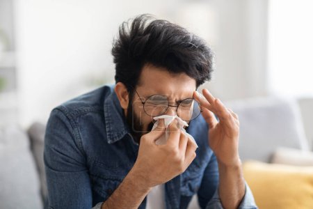Téléchargez les photos : Jeune Indien malade qui souffle du nez dans du papier tout en étant assis sur un canapé à la maison, un homme de l'Est du millénaire souffrant de grippe saisonnière ou de rhume, ayant une rhinite ou une réaction allergique, gros plan - en image libre de droit