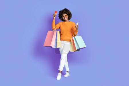Photo for Cool stylish happy smiling young black woman with bushy hair wearing sunglasses holding colorful shopping bags, smartphone, bank card, posing over purple studio background. Unlimited shopping - Royalty Free Image