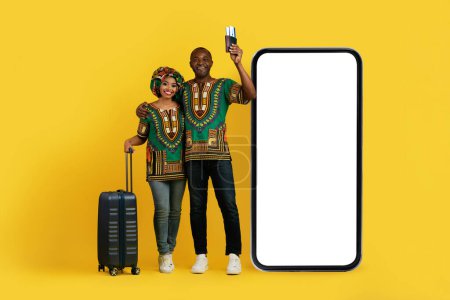Téléchargez les photos : Heureux couple noir joyeux portant des costumes africains traditionnels voyageurs en vacances, portant des bagages, tenant un passeport, debout par un énorme téléphone avec écran blanc, réservation de billets en ligne - en image libre de droit