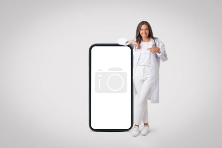 Téléchargez les photos : Femme médecin senior heureuse positive en uniforme blanc posant et pointant vers un grand téléphone portable avec écran blanc vide, fond de studio gris, maquette. Concept de télémédecine - en image libre de droit