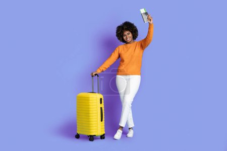 Téléchargez les photos : Joyeux millénium élégant afro-américaine voyageant à l'étranger, ayant des vacances, montrant des billets et un passeport, portant des bagages, isolé sur fond de studio violet, espace de copie - en image libre de droit