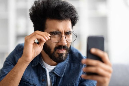 Téléchargez les photos : Concept des problèmes de vue. Jeune homme indien dans les lunettes de vue regardant l'écran de Smartphone et plissant, gars oriental essayant de lire le message, souffrant d'asstigmatisme et de mauvaise vision, gros plan - en image libre de droit