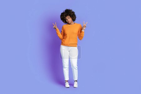 Téléchargez les photos : Cool jolie insouciante élégante jeune femme noire aux cheveux touffus portant tenue décontractée montrant geste de paix avec les deux mains, fond violet studio, pleine longueur, espace de copie - en image libre de droit