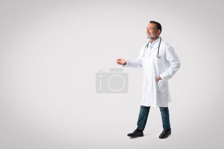 Téléchargez les photos : Souriant médecin vieil homme confiant en manteau blanc avec stéthoscope va, saluant et montre la main à l'espace vide sur fond de studio gris. Soins de santé, service médical, traitement de la maladie du patient - en image libre de droit