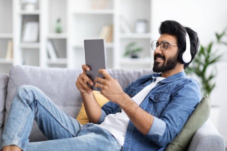 Photo for Handsome Indian Man In Wireless Headphones Using Digital Tablet At Home, Smiling Eastern Male Browsing Internet Or Shopping Online With Modern Gadget While Relaxing On Couch In Living Room, Side View - Royalty Free Image