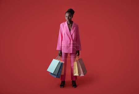 Photo for Fashion Shot Of Young Black Stylish Woman With Paper Shopping Bags In Hands Standing Over Red Background In Studio, Full Length Of African American Female Model Wearing Pink Suit, Copy Space - Royalty Free Image