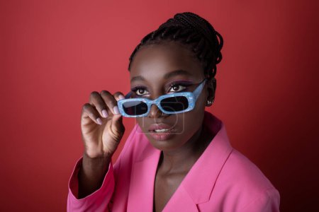 Foto de Primer plano de hermosa modelo de moda femenina negra con gafas de sol elegantes, retrato de mujer afroamericana joven con chaqueta de chaqueta rosa y gafas de moda posando sobre fondo rojo - Imagen libre de derechos