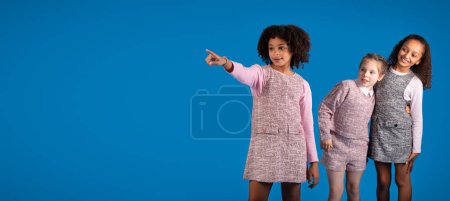 Téléchargez les photos : Inspiré heureux multiracial petites amies dans les doigts pointant occasionnels à l'espace de copie, amusez-vous isolé sur fond bleu, studio. Éducation ensemble, émotions des enfants, vente et offre - en image libre de droit