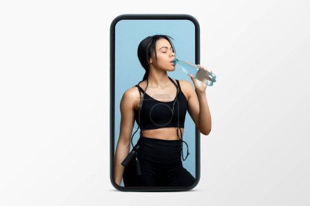 Photo for Thirsty millennial black woman in sportswear with jump rope drinks water from bottle on phone screen isolated on white studio background. Sports blog, app for workout and body care - Royalty Free Image