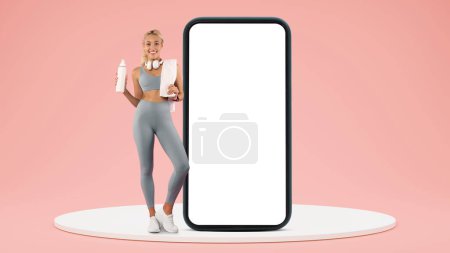 Photo for Positive young caucasian woman in sportswear with towel resting with water bottle on platform near smartphone screen isolated on pink studio background. Fit, aqua balance, app for sports - Royalty Free Image