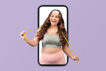 Photo for Smiling young caucasian woman plus size in sportswear and measuring tape on smartphone screen isolated on violet studio background. Body care, sports, app for weight loss and fitness - Royalty Free Image