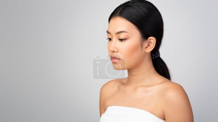 Photo for Bodycare Offer. Gorgeous Asian Woman Posing Near Copy Space, Standing Wrapped In Towel And Looking Aside On Gray Studio Background. Beauty And Pampering Advertisement Concept. Panorama - Royalty Free Image