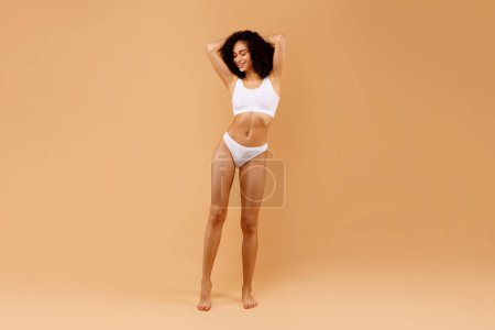 Téléchargez les photos : Plan complet de jeune femme noire sensuelle posant en sous-vêtements avec les mains derrière la tête sur fond de studio beige, montrant la forme parfaite du corps, espace de copie - en image libre de droit
