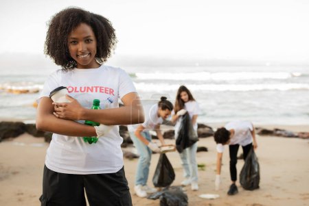 Foto de Sonriente joven afroamericana dama celebrar botellas y personas multirraciales voluntarios en camisetas con bolsas de basura limpiar la basura en la playa de mar, al aire libre. Conservación del medio ambiente, eco - Imagen libre de derechos