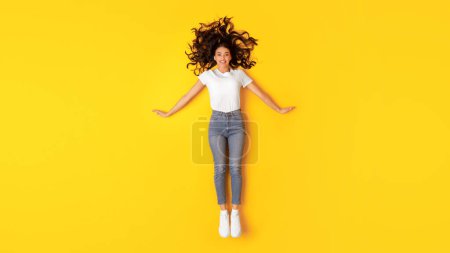 Téléchargez les photos : Offre beauté. Jolie jeune femme allongée sur le sol posant souriant à la caméra sur fond jaune studio, vue de dessus. Prise de vue pleine longueur d'une femme heureuse portant un t-shirt blanc et un jean maigre - en image libre de droit