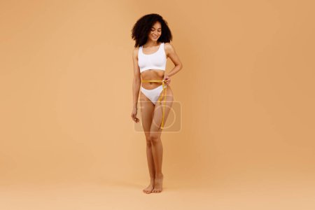 Photo for Healthy lifestyle concept. Fit black lady in underwear measuring her waistline with measure tape, posing over beige background, woman with sporty body checking measures, full length - Royalty Free Image