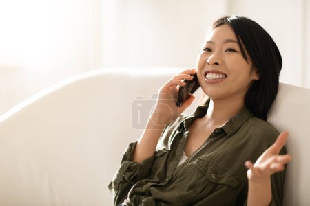 Téléchargez les photos : Heureuse joyeuse souriante belle femme asiatique millénaire dans des tenues décontractées confortables assis sur le canapé, ayant une conversation téléphonique avec un ami ou un amant à la maison, regardant l'espace de copie et riant, gros plan - en image libre de droit