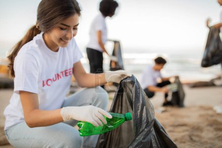 Téléchargez les photos : Heureux les jeunes bénévoles et dame asiatique dans des gants avec sac poubelle nettoyer les ordures sur la plage de la mer, en plein air. Conservation de l'environnement, protection de la planète, Jour de la Terre et pollution des océans - en image libre de droit