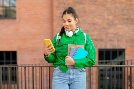 Téléchargez les photos : Joyeuse étudiante de race mixte décontractée avec sac à dos tenant des livres et utilisant un smartphone, debout près de l'université en plein air. Gadget et application pour l'étude, la connaissance, concept d'éducation - en image libre de droit