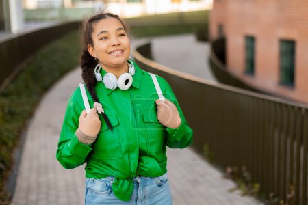 Téléchargez les photos : Jeune fille étudiante mixte à la mode marchant sur le campus universitaire, portant un sac à dos et un casque au cou, se reposant après avoir étudié à l'extérieur, regardant ailleurs et souriant, copiant l'espace - en image libre de droit