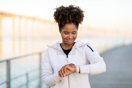 Téléchargez les photos : Femme noire souriante regardant Smartwatch, Vérification de l'activité sportive après l'entraînement en plein air, Jeune joggeuse afro-américaine contrôlant l'endurance, Regarder le temps sur la montre, Copier l'espace - en image libre de droit