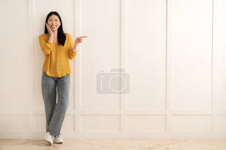 Téléchargez les photos : Incroyable heureux joyeux jolie jeune femme asiatique portant tenue décontractée élégant debout par mur vide à la maison, pointant vers l'espace de copie pour la publicité, toucher son visage, pleine longueur - en image libre de droit