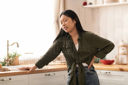 Photo for Exhausted young asian woman in casual suffering from tiredness, standing next to table in kitchen, holding her lower back, copy space. Gastrointestinal disorders, appendicitis, backache concept - Royalty Free Image