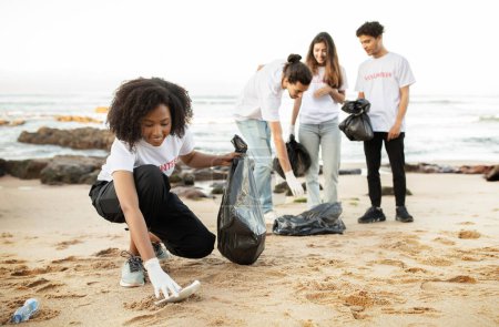 Téléchargez les photos : Heureux les jeunes multiethniques bénévoles en t-shirts avec des paquets de déchets nettoyer les ordures, bouteilles en plastique sur la plage de la mer, en plein air. Conservation de l'environnement, protection de l'environnement - en image libre de droit