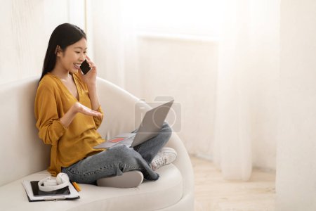 Happy pretty young asian woman in casual sitting on couch at home, using laptop, bank card, talking on phone, have conversation with customer service representative, shopping online, copy space