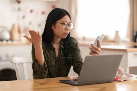 Téléchargez les photos : Irrité confus jeune asiatique dame portant tenue et lunettes assis au bureau de la cuisine en face de l'ordinateur portable, travailler de la maison, regarder l'écran du téléphone et des gestes, eu de mauvaises nouvelles, copier l'espace - en image libre de droit