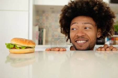 Téléchargez les photos : Tentation de la malbouffe. Homme noir affamé drôle regardant hamburger savoureux placé sur la table dans la cuisine moderne à la maison. Nutrition malsaine et Cheat fringales de repas pendant la perte de poids Diet Concept - en image libre de droit
