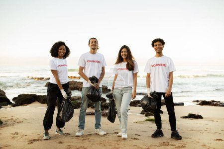 Téléchargez les photos : Souriant jeunes différentes personnes bénévoles en t-shirts et gants en caoutchouc avec des paquets d'ordures nettoyer les ordures sur la plage de la mer, en plein air. Conservation de l'environnement, protection de la planète, Jour de la Terre - en image libre de droit