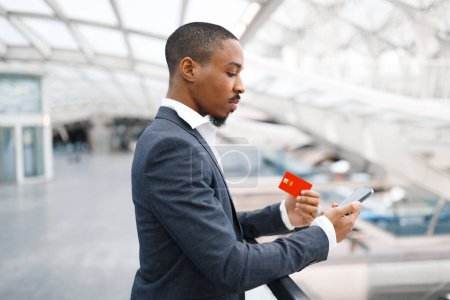 Photo for Young Black Businessman In Airport Using Smartphone And Credit Card, Handsome Millennial African American Man In Suit Making Online Payments While Waiting Flight Departure At Terminal, Side View - Royalty Free Image