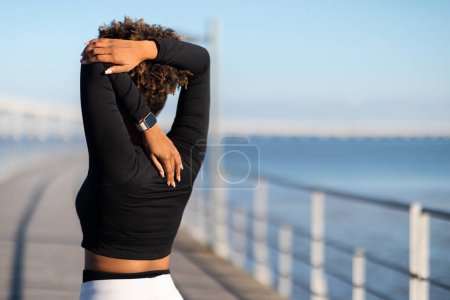Foto de Mujer negra irreconocible estirándose al aire libre, preparándose para el ejercicio físico exterior, atleta joven afroamericana usando smartwatch, haciendo rutina de calentamiento en muelle cerca del mar, espacio de copia - Imagen libre de derechos