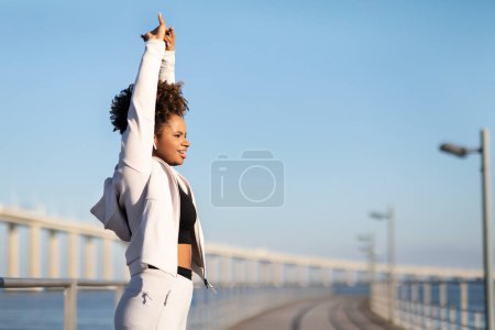 Téléchargez les photos : Belle jeune femme noire faisant des exercices d'étirement pendant l'entraînement en plein air, Souriante athlète afro-américaine levant les mains, Réchauffer les muscles pendant l'entraînement à l'extérieur, Copier l'espace - en image libre de droit
