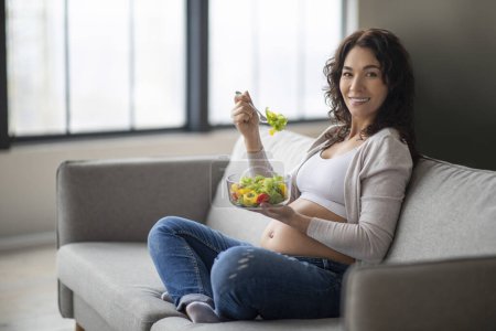 Photo for Nutrition Needs During Pregnancy. Pregnant Woman Eating Fresh Vegetable Salad At Home, Smiling Young Expectant Mother Sitting On Couch And Enjoying Healthy Meal, Having Vitamin Food, Copy Space - Royalty Free Image