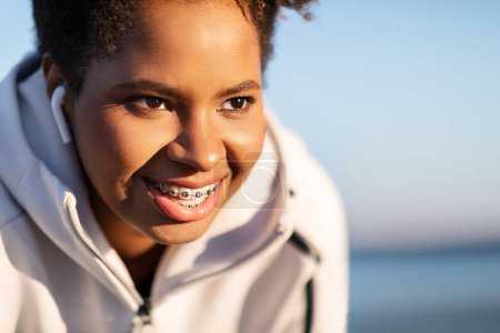Photo for Closeup Portrait Of Smiling Young Black Female Athlete Resting Outdoors After Training, Motivated Sporty African American Woman Wearing Wireless Earphones And Dental Braces Looking Away And Smiling - Royalty Free Image