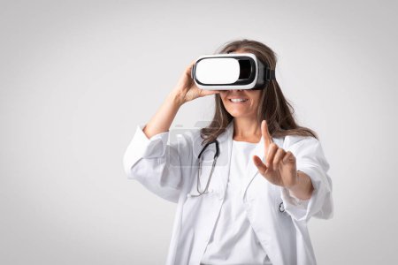Photo for VR medicine concept. Senior woman doctor using modern virtual reality headset for medical purposes, standing over light studio background, free space - Royalty Free Image